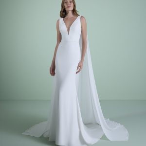 Bröllopsklänning Amphitrite med figurnära siluett, djup V-ringning och lång axelkappa – modern och elegant design för stilrena bröllop.