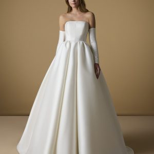 Brud i elegant axelbandslös Mephis-bröllopsklänning från Nicole Milano i mikado med A-linjesiluett och avtagbara ärmar, stående mot beige bakgrund