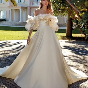 Brud i Pronovias Regina-klänning med volangprydd off-shoulder överdel och A-linjesiluett i mikado, poserar framför villa i trädgårdsmiljö.